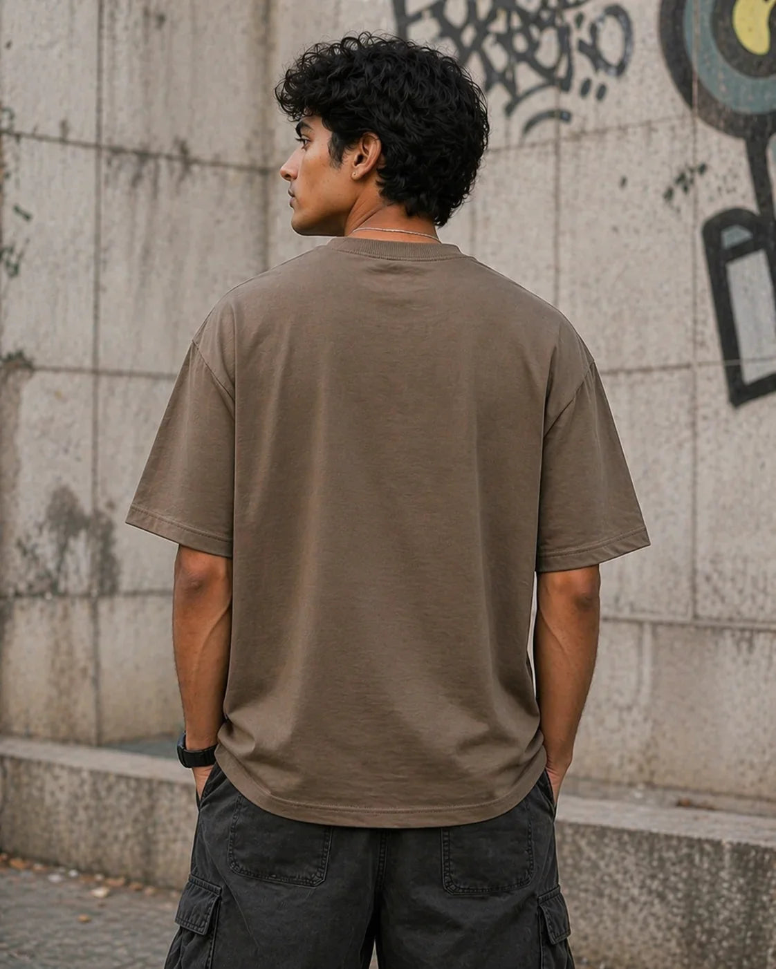 Man wearing oversized brown t-shirt and cargo shorts standing by graffiti wall outdoors