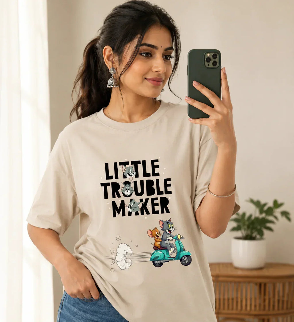 Woman wearing oversized beige t-shirt featuring cartoon cat and mouse riding scooter, text 'Little Trouble Maker'.