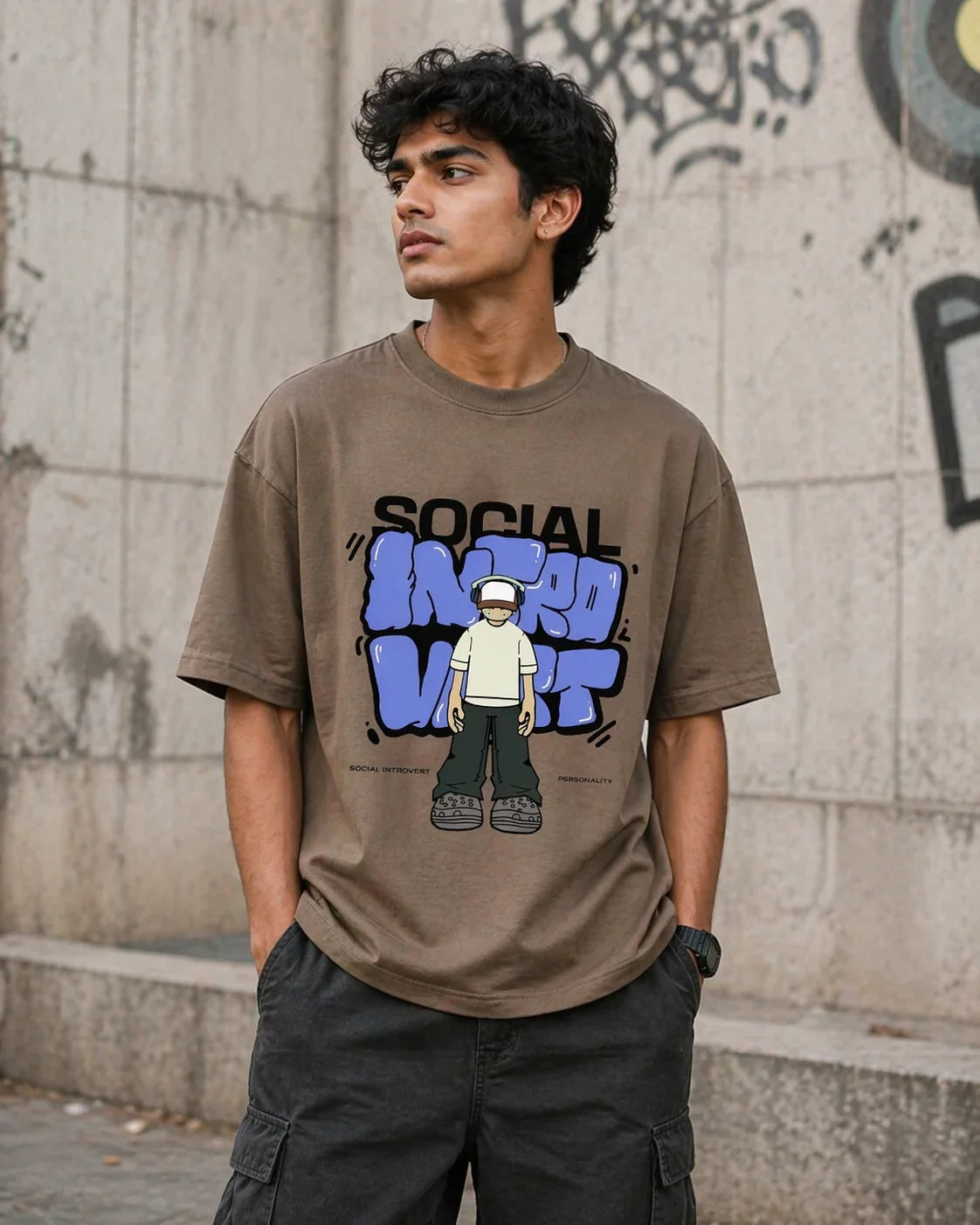 Man wearing oversized brown anime graphic t-shirt with 'Social Introvert' print, urban wall background