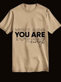 Beige oversized t-shirt with 'YOU ARE enough' motivational text print, laid flat on black background