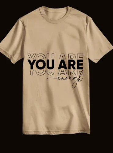 Beige oversized t-shirt with 'YOU ARE enough' motivational text print, laid flat on black background
